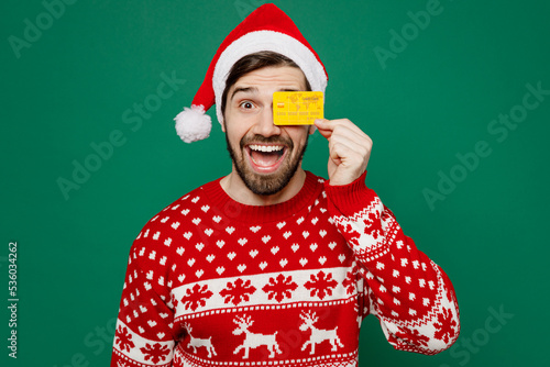 Merry young man 20s in red warm knitted sweater Santa hat posing hold in hand cover eye with credit bank card isolated on plain dark green background studio New Year 2023 holiday celebration concept