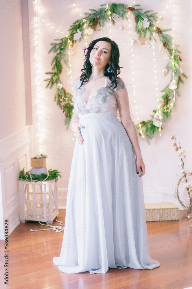 Young pregnant woman with dark hair in airy dress in a room decorated with pine needles and sparkling garlands for Christmas. Christmas mood . Pregnancy Young pregnant woman with dark hair in airy dress in a room decorated with pine needles and sparkling garlands for Christmas. Christmas mood . Pregnancy