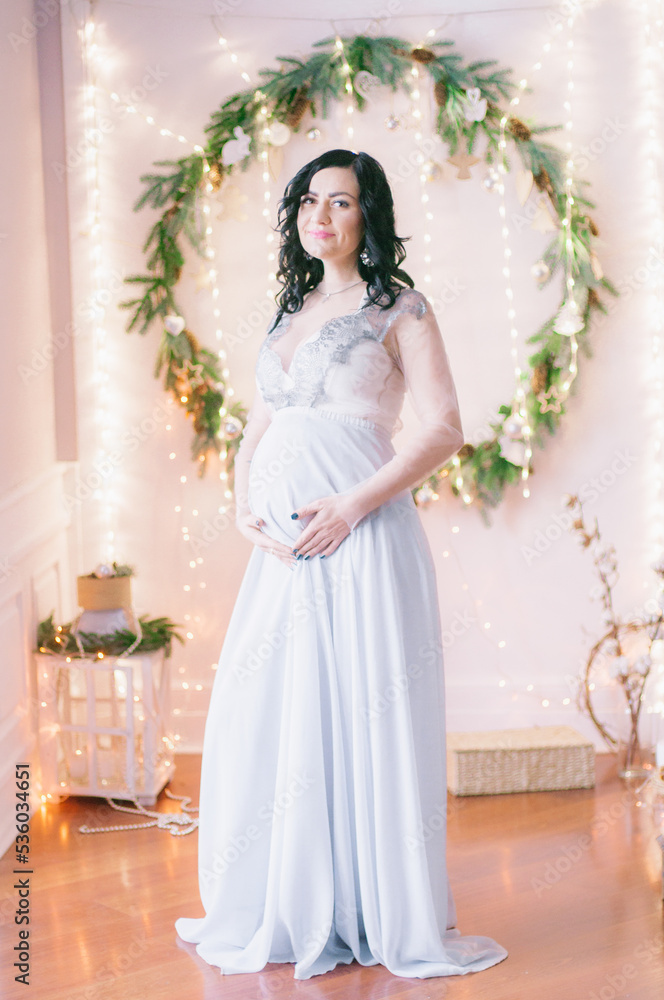 Young pregnant woman with dark hair in airy dress in a room decorated with pine needles and sparkling garlands for Christmas. Christmas mood . Pregnancy Young pregnant woman with dark hair in airy dress in a room decorated with pine needles and sparkling garlands for Christmas. Christmas mood . Pregnancy