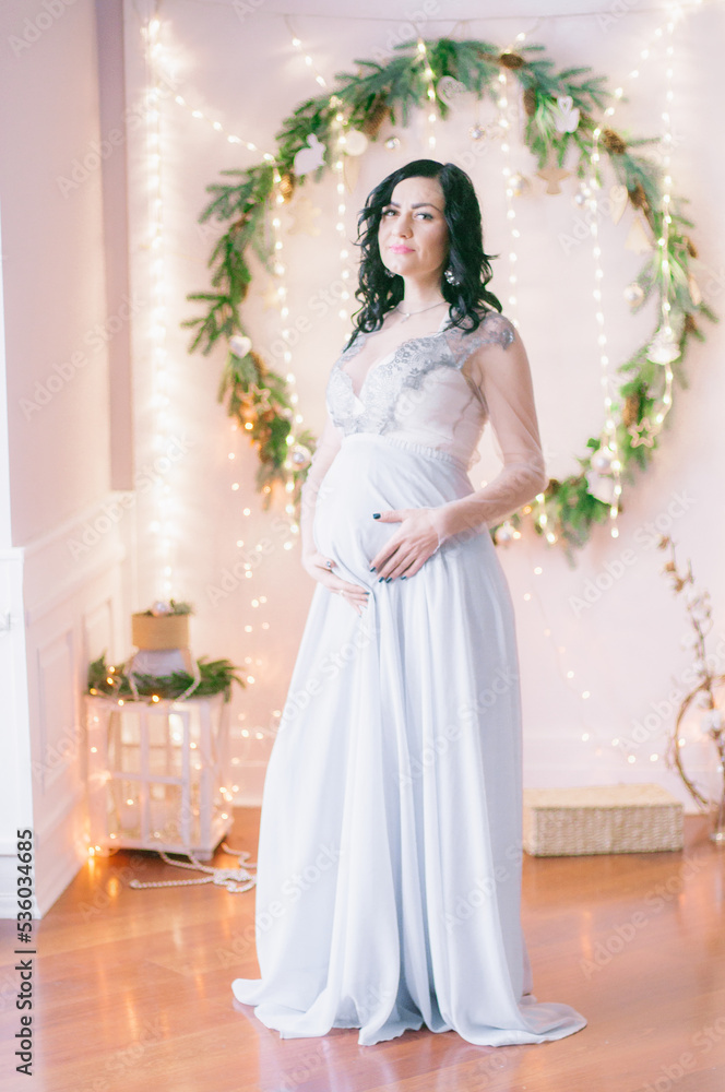 Young pregnant woman with dark hair in airy dress in a room decorated with pine needles and sparkling garlands for Christmas. Christmas mood . Pregnancy Young pregnant woman with dark hair in airy dress in a room decorated with pine needles and sparkling garlands for Christmas. Christmas mood . Pregnancy