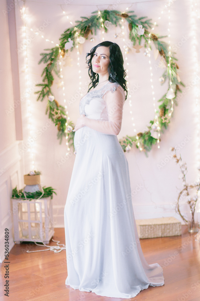Young pregnant woman with dark hair in airy dress in a room decorated with pine needles and sparkling garlands for Christmas. Christmas mood . Pregnancy Young pregnant woman with dark hair in airy dress in a room decorated with pine needles and sparkling garlands for Christmas. Christmas mood . Pregnancy
