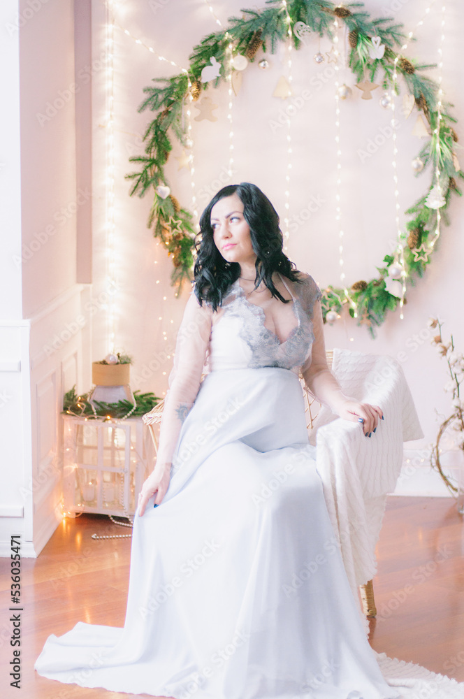 Young pregnant woman with dark hair in airy dress in a room decorated with pine needles and sparkling garlands for Christmas. Christmas mood . Pregnancy Young pregnant woman with dark hair in airy dress in a room decorated with pine needles and sparkling garlands for Christmas. Christmas mood . Pregnancy