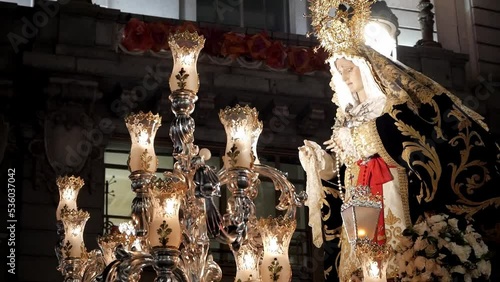 Close-up of Virgin Mary figure on carriage float during Holy Week celebrations in Madrid, Spain. Procession is rocking the figure lit up with lantern at night. Building with Eurpean flag behind.