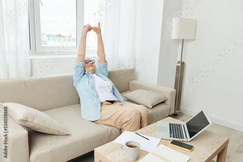 a joyful elderly lady is resting sitting on a comfortable sofa in her apartment, relaxing, raising her hands up, stretching after a working day with a laptop and notebook standing on the table