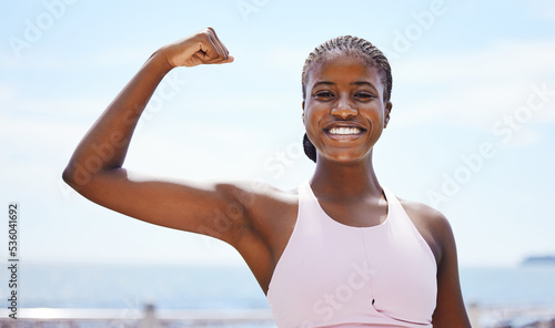 Fitness, portrait and black woman flexing muscle at the beach in summer after strength training, exercise and workout. Smile, motivation and happy girl outdoors with healthy and strong arm biceps photo