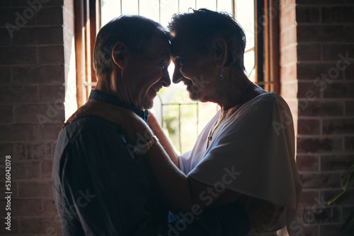 Love, dance and elderly couple hug in living room window, romantic and bonding in their home together. Romance, retirement and sweet seniors sharing a romantic moment, dancing and embracing in house