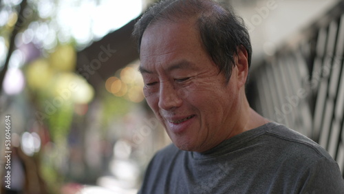 Happy senior man using cellphone standing outdoors in city street. Portrait older Asian American person typing message on smartphone device smiling