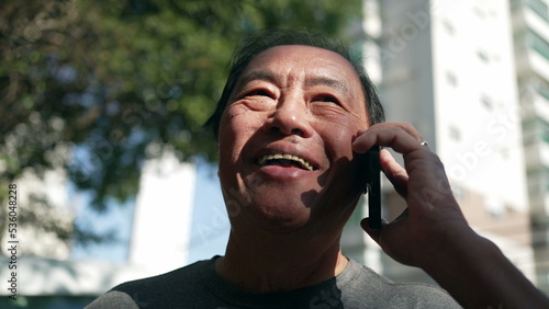 Smiling happy middle aged Asian man speaking on cellphone in street. Happy Person holding smartphone in ear. Portrait guy in conversation