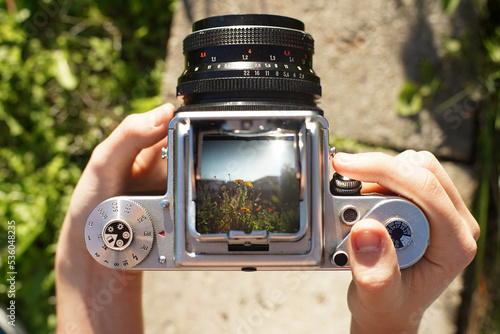 A photographer holding a vintage analog photo camera focusing ajusting taking pictures outside looking through a large square format viewfinder mechanical settings solid metal cogs gagarden background photo