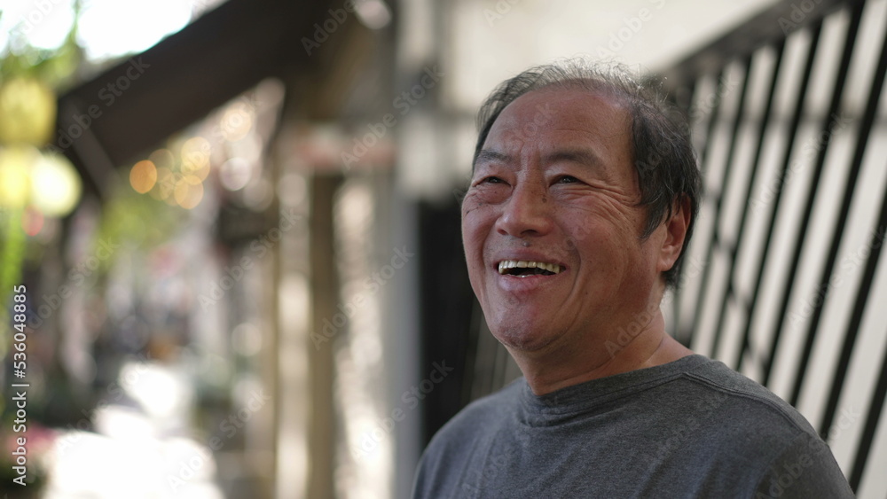 Fototapeta premium A happy Senior Asian man standing in street sidewalk smiling. Portrait of a middle aged older joyful person in daylight. Authentic real life smile