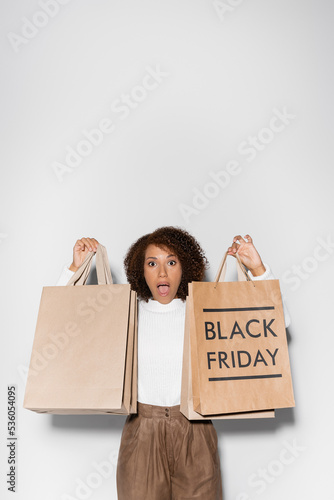 shocked african american woman in autumnal outfit holding shopping bags with black friday lettering on grey. © LIGHTFIELD STUDIOS