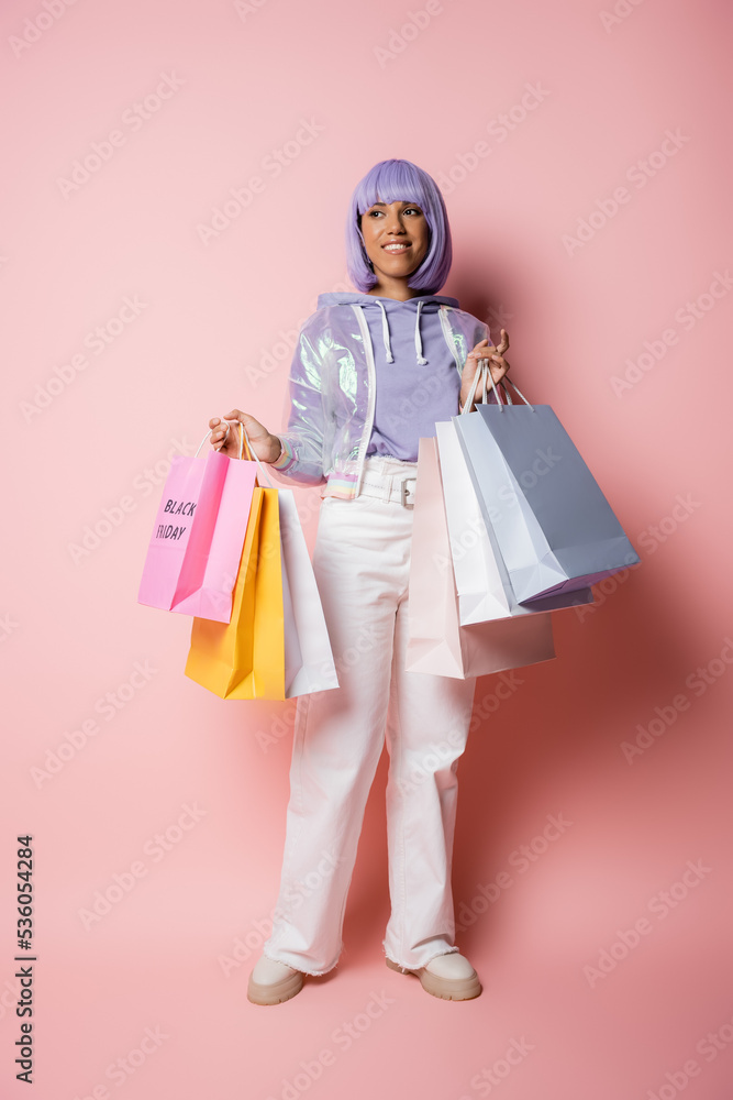 full length of smiling african american woman with purple hair holding shopping bags with black friday lettering on pink.
