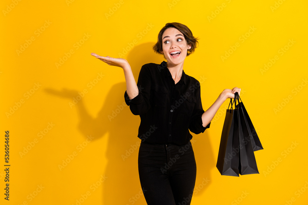 Photo of excited thoughtful lady wear black shirt rising arm empty space shoppers isolated yellow color background Photo of excited thoughtful lady wear black shirt rising arm empty space shoppers isolated yellow color background