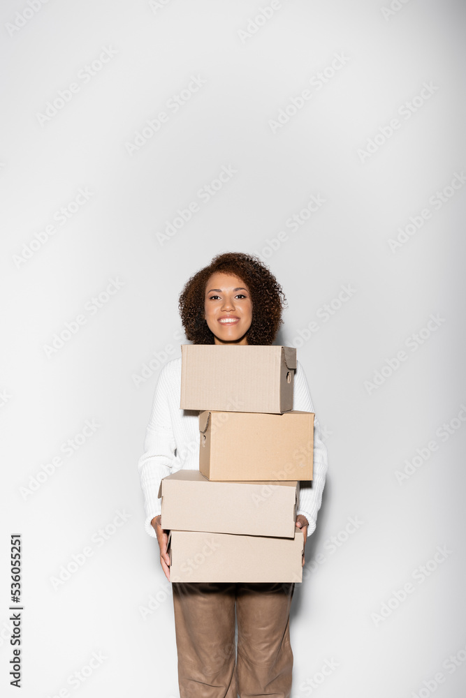 delighted african american woman with curly hair holding delivery boxes on grey. delighted african american woman with curly hair holding delivery boxes on grey.
