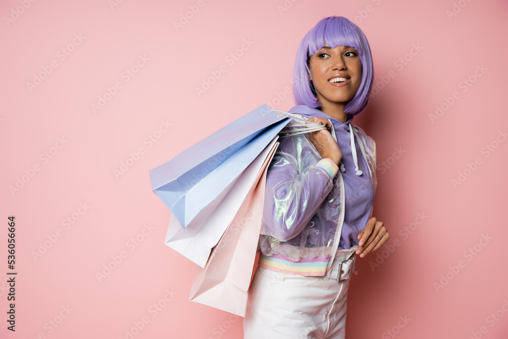 joyful african american woman with purple hair standing in transparent jacket with shopping bags on pink.