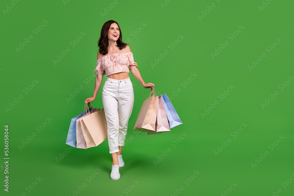 Full length photo of excited pretty lady wear print top walking holding shoppers looking empty space isolated green color background