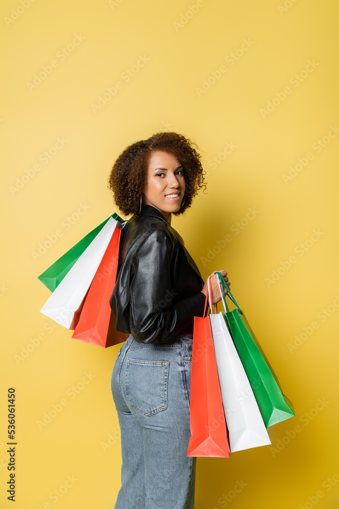 pleased african american woman in stylish leather jacket and jeans holding black friday shopping bags on yellow.
