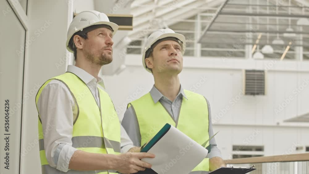 Waist up slowmo of two mid aged Caucasian engineers in hard hats and reflective vests looking at building plans while standing at construction site indoors having discussion Waist up slowmo of two mid aged Caucasian engineers in hard hats and reflective vests looking at building plans while standing at construction site indoors having discussion