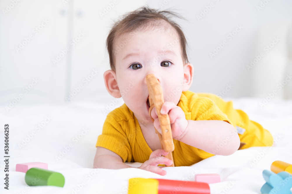 healthy asian baby girl in yellow bodysuit plays with wooden toy teether for teeth on white bedding healthy asian baby girl in yellow bodysuit plays with wooden toy teether for teeth on white bedding