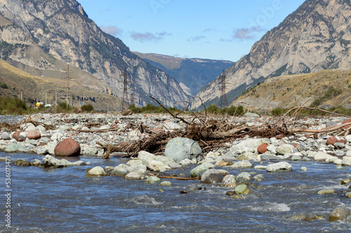 Mountain stream in September. Blue and grey water, white and brown stones, green and brown rocky mountain slopes, blue sky with clouds