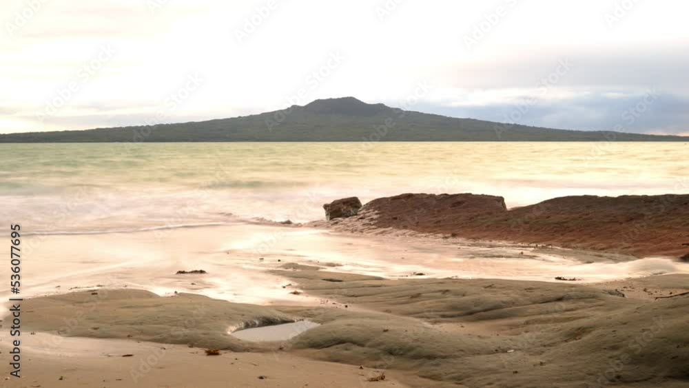 Time lapse of the sun rising behind Rangitoto volcano, viewed from a rocky beach across the turquoise ocean Time lapse of the sun rising behind Rangitoto volcano, viewed from a rocky beach across the turquoise ocean