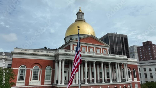 Slow aerial pass of American flag in front of Massachusetts state house. Historical government capitol building in Boston. American government, democracy, federal system theme.