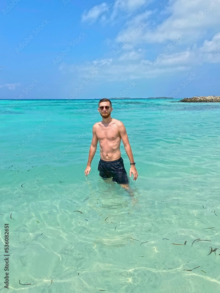 Handsome young man standing on the beach in the island of Maldives, shirtless in boxer shorts, showing off his muscular body. Incredibly beautiful turquoise ocean Handsome young man standing on the beach in the island of Maldives, shirtless in boxer shorts, showing off his muscular body. Incredibly beautiful turquoise ocean