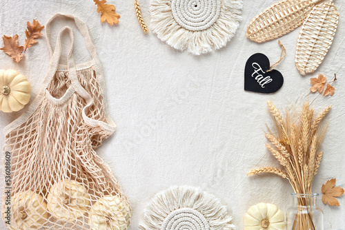 Off white textile background with natural Fall decorations. Flat lay, top view. Pumpkins in net bag, dry leaves, macrame pads, wattle leaves, wheat ears. Autumn text, word on black wooden heart.