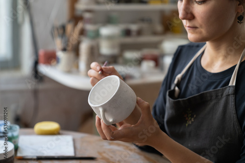 Craftswoman creates new masterpiece in work studio making sketch of future pattern on white mug. Lady master wants to decorate kitchen with painted clay dish using pencil to draw interesting pattern photo