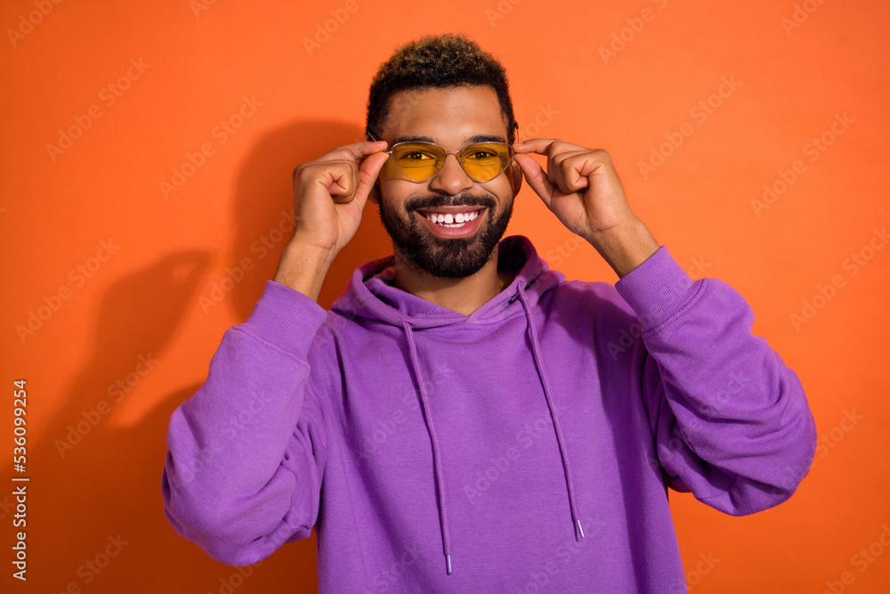 Portrait of positive nice young man toothy beaming smile hands touch sunglass isolated on orange color background Portrait of positive nice young man toothy beaming smile hands touch sunglass isolated on orange color background