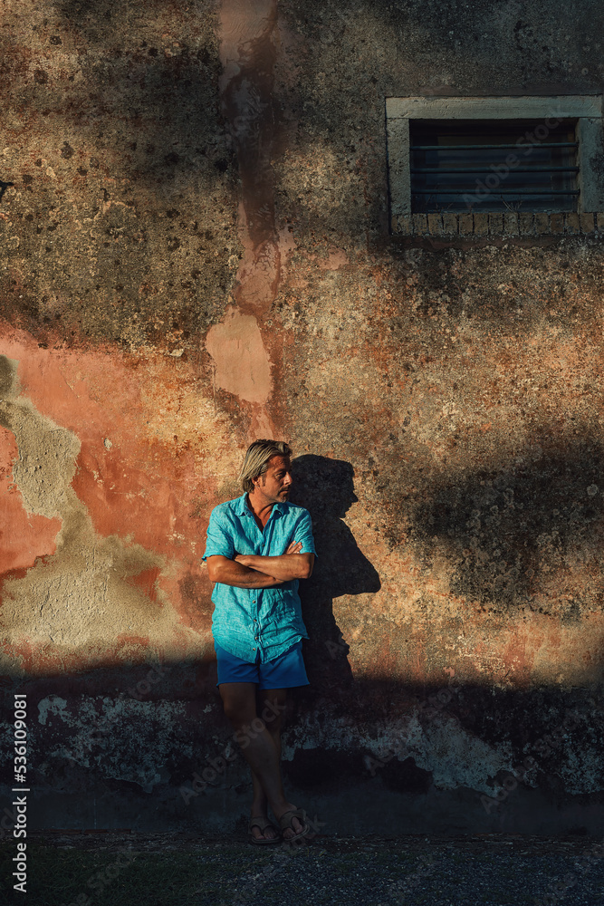 Blond man in blue shirt and swim shorts stands against weathered pink plaster wall in late afternoon sunlight. Blond man in blue shirt and swim shorts stands against weathered pink plaster wall in late afternoon sunlight.
