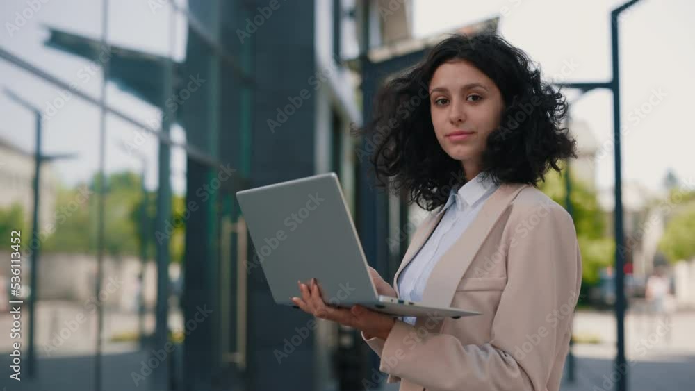 Portrait of the beautiful curly smiling young businesswoman in formal outfit standing near the business centre looking at the camera holding and using laptop. People and technology business concept Portrait of the beautiful curly smiling young businesswoman in formal outfit standing near the business centre looking at the camera holding and using laptop. People and technology business concept