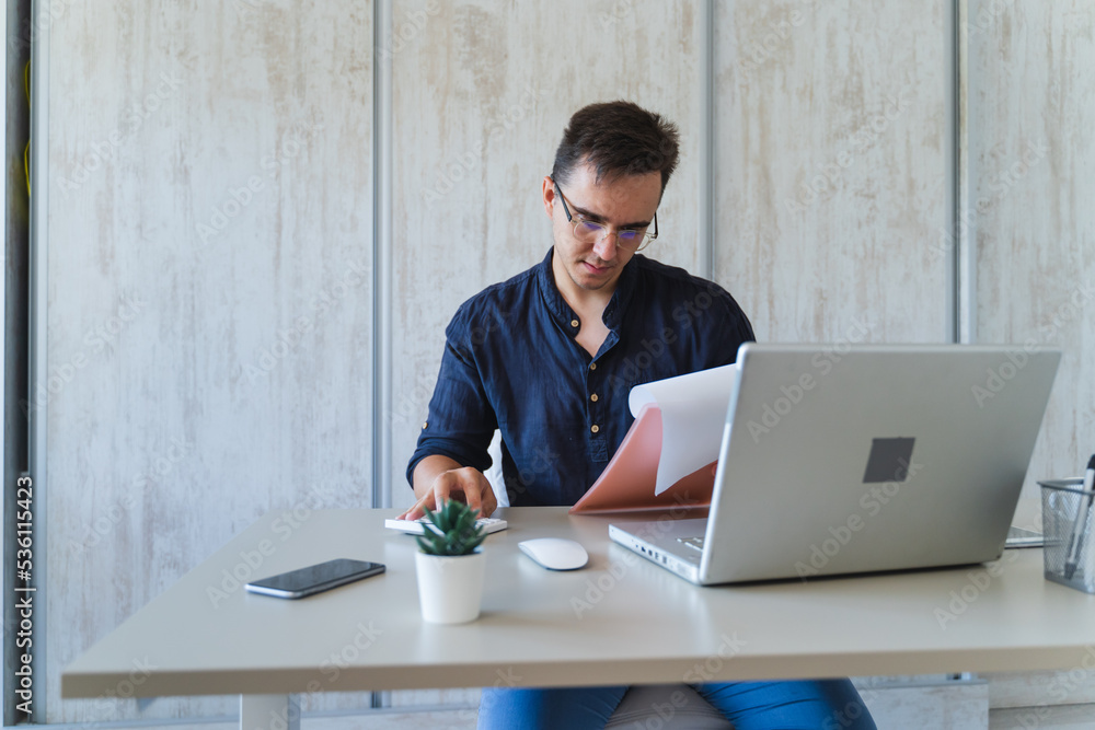 A young guy doing business work in his home office during the day while talking on phone using laptop and his mobile phone A young guy doing business work in his home office during the day while talking on phone using laptop and his mobile phone