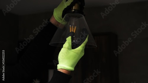  A man in gloves unscrews a lamp from a parton near a hanging ceiling lamp. Unscrewing a burnt out light bulb, replacing the bulb with an electrician in slow motion. The concept of saving electricity  photo