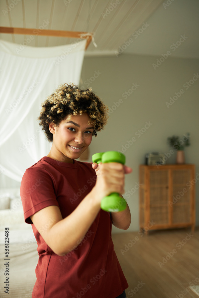 Vertical indoor image of cute funny young woman with afro hairstyle standing in bedroom with green dumbbells in hands pretending like boxing, looking at camera with smile. Fitness at home Vertical indoor image of cute funny young woman with afro hairstyle standing in bedroom with green dumbbells in hands pretending like boxing, looking at camera with smile. Fitness at home
