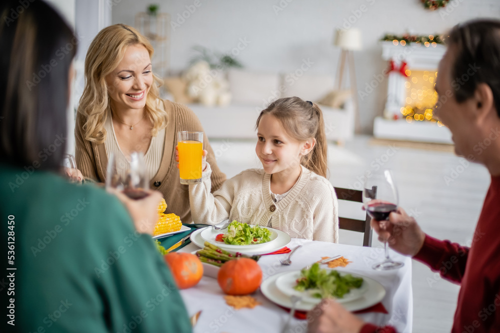 Smiling girl holding orange juice near mother and blurred grandparents during thanksgiving celebration