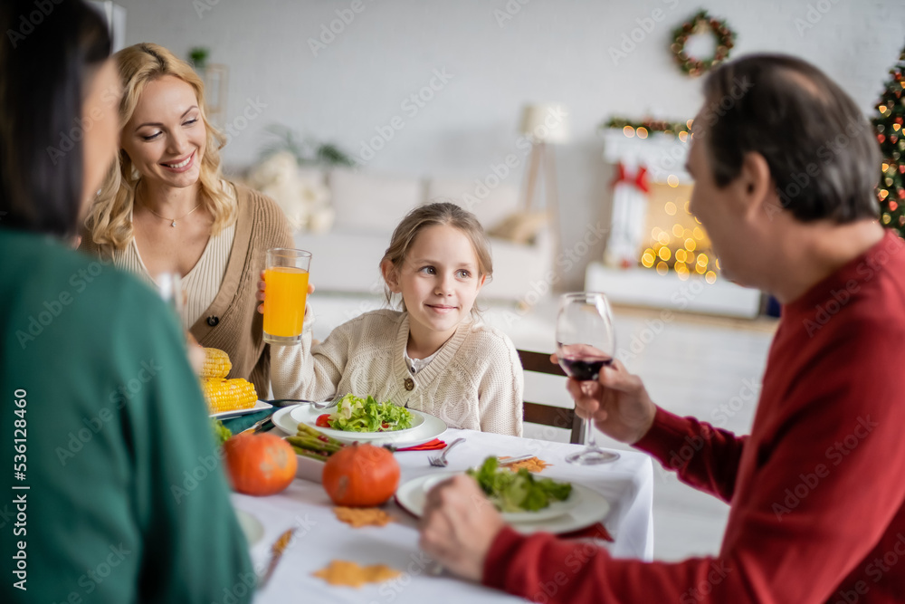 Smiling kid with orange juice looking at blurred grandparent during thanksgiving celebration at home