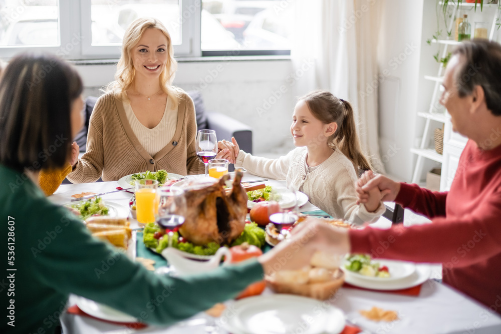 Smiling woman looking at camera while family holding hands near thanksgiving dinner at home