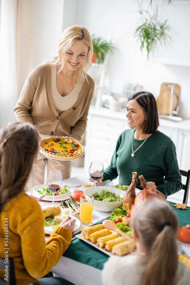 Positive woman holding tasty thanksgiving pie near multiethnic mother and children at home