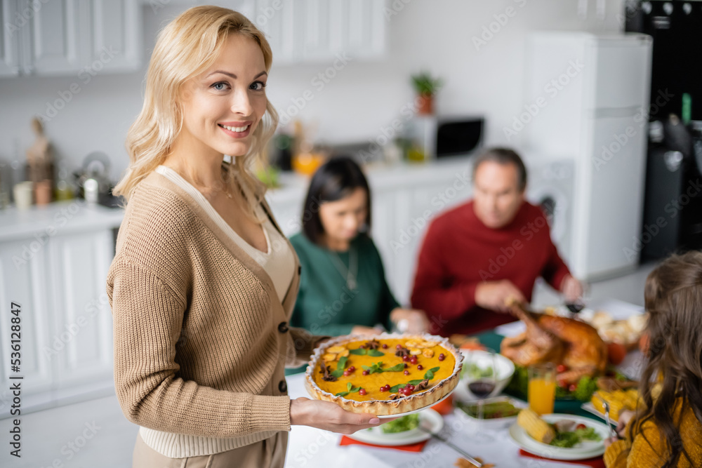 Smiling woman holding thanksgiving pie and looking at camera near blurred family at home