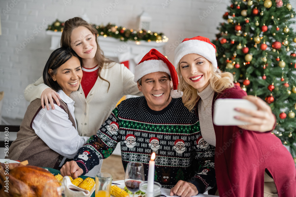 smiling woman in santa hat taking selfie with multiethnic family near festive dinner and christmas tree smiling woman in santa hat taking selfie with multiethnic family near festive dinner and christmas tree