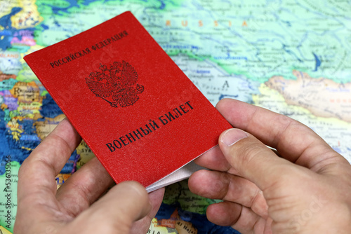 Russian military ID in male hands on background of map of Russia and Europe, translation of inscription: Military ID card, Russian Federation. Concept of mobilization and war