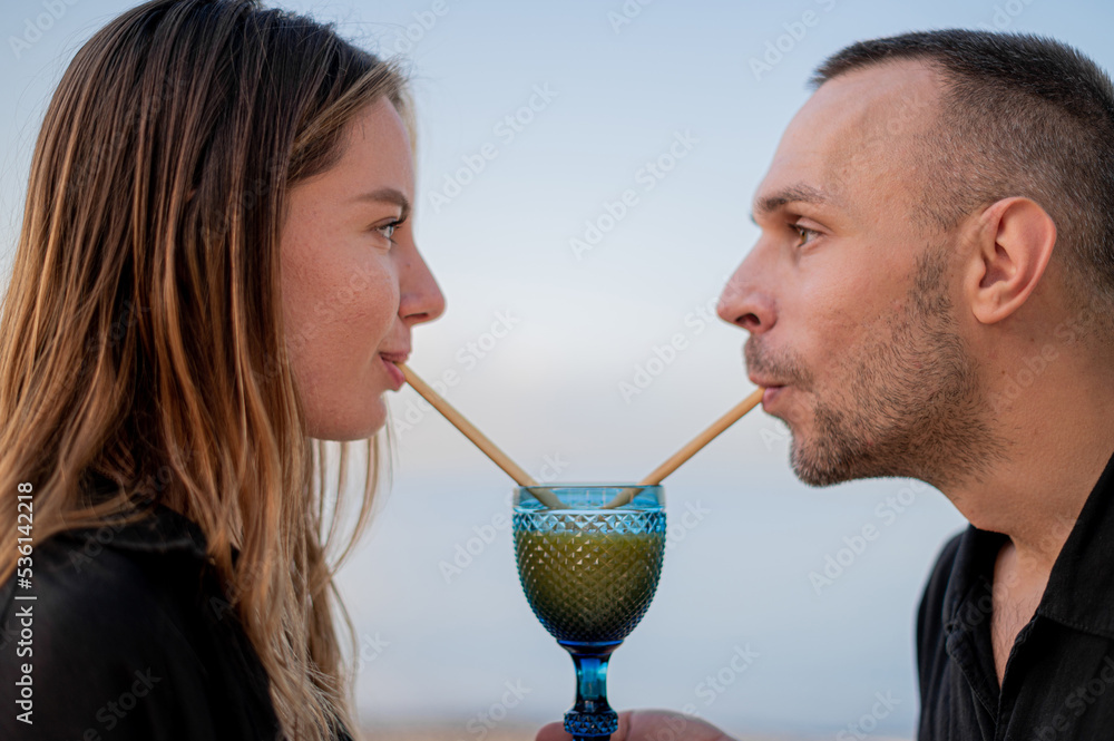 Fototapeta premium Young couple sitting on a beach at sunset and looking at each drinking from one glass with a drinking straw