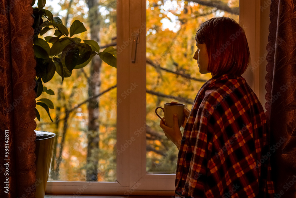 Autumn coffee. A beautiful girl with a cup of coffee in her hands sits and looks out the window. Cozy autumn composition. Copy space.