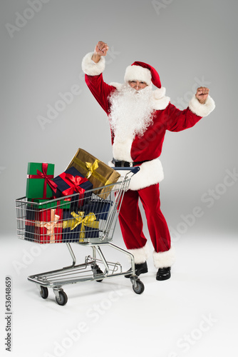 Full length of excited santa claus standing near shopping cart with gifts on grey background. © LIGHTFIELD STUDIOS