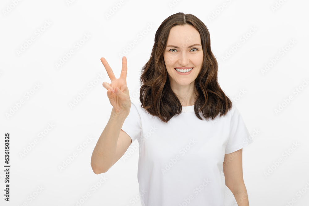 brunette smiling woman in white T-shirt shows peace gesture on white background with fingers brunette smiling woman in white T-shirt shows peace gesture on white background with fingers