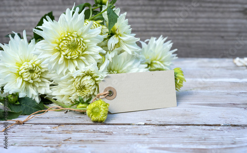 A bouquet of white dahlia flowers and a card with copy space are on a wooden background.