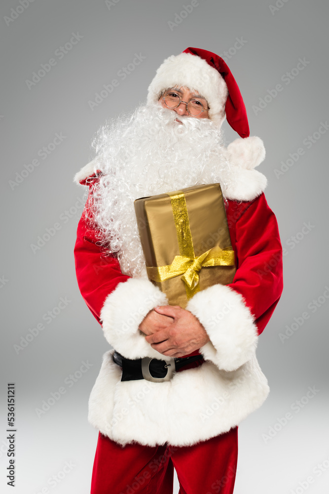 Santa claus in hat and eyeglasses holding present and looking at camera isolated on grey. Santa claus in hat and eyeglasses holding present and looking at camera isolated on grey.