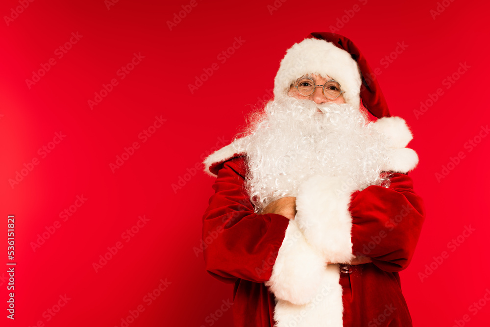 Bearded santa claus crossing arms and looking at camera on red background.