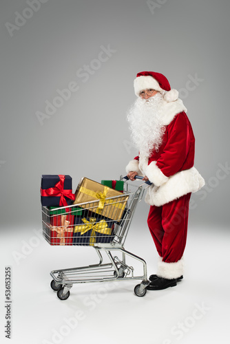 Full length of santa claus in costume and eyeglasses standing near shopping cart with gifts on grey background. © LIGHTFIELD STUDIOS